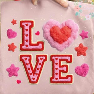 Puede incluir: Un artículo de tela rosa con la palabra "LOVE" bordada en rojo y rosa. La "O" tiene forma de corazón esponjoso, y el diseño incluye estrellas y corazones. La imagen tiene una marca de agua de archivo digital.