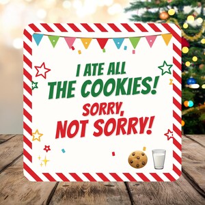 May include: A square sign with a red and white diagonal striped border. The white centre features colourful bunting, stars, and confetti. Text reads: I ATE ALL THE COOKIES! SORRY, NOT SORRY! A chocolate chip cookie and a glass of milk are at the bottom.