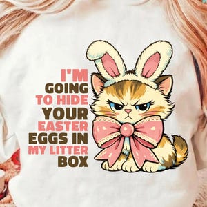 May include: White t-shirt featuring a cartoon cat wearing bunny ears and a pink bow. The cat has an angry expression and the text reads "I'M GOING TO HIDE YOUR EASTER EGGS IN MY LITTER BOX."