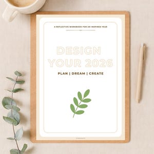 Op de afbeelding: Een werkboek getiteld "DESIGN YOUR 2026" met de tekst "PLAN | DREAM | CREATE" op een houten klembord. Een kop koffie en een pen liggen naast het werkboek. Een takje groene bladeren staat onderaan het werkboek.
