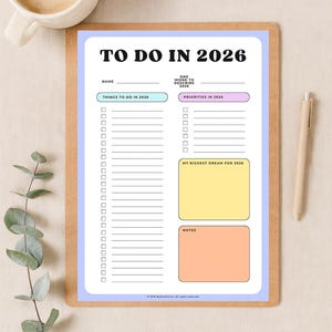 Op de afbeelding: Een planner getiteld "TO DO IN 2026" met secties voor taken, prioriteiten en dromen. De planner heeft een lichtblauwe rand en een houten klembordachterkant. Een pen en een kopje koffie liggen naast de planner.