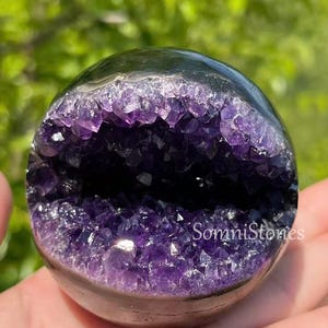 May include: A dark, polished sphere with a cavity filled with vibrant purple amethyst crystals. The crystals sparkle within the dark, smooth exterior. The text "SomniStones" is visible on the sphere.