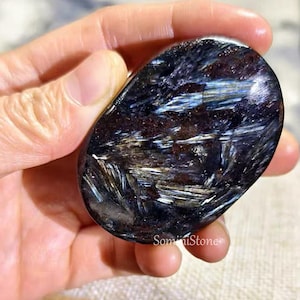 Pode incluir: Uma pedra preciosa polida, de formato oval, com um esquema de cores azul escuro e castanho. A pedra apresenta um padrão único de riscas azuis claras e brancas, criando uma aparência texturizada. A pedra preciosa é segurada em uma mão.