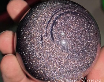 May include: A dark, glittery sphere with a swirling design. The sphere is held in a hand, and the surface sparkles with flecks of gold and silver. The sphere has a polished, smooth surface. The text "SomniStones" is visible.