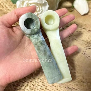 May include: Two stone pipes, one green and one pale yellow, are held in a hand. The green pipe has a mottled pattern, while the yellow pipe is solid. The text "SomniStones" is visible on the image.