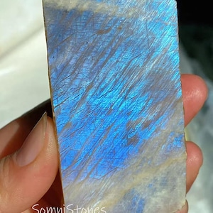 以下が含まれることがあります： 鮮やかな青色の虹色を帯びた、研磨された長方形の宝石。石は自然で粗い質感で、白とグレーの縞模様があります。「SomniStones」の文字が石の下部に表示されています。