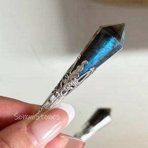 May include: A labradorite crystal pendant with a silver filigree setting. The crystal has a dark base with iridescent blue flashes. The silver setting has an ornate, openwork design. The text "SomniStones" is visible.