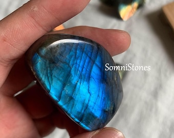 May include: A polished labradorite stone with a vibrant blue and iridescent sheen. The stone is held in a hand, showcasing its smooth, rounded shape and natural beauty. The text "SomniStones" is visible on the right side.