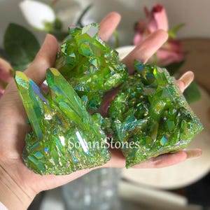Può includere: Tre gruppi di cristalli di quarzo aura verde. I cristalli hanno una superficie scintillante e iridescente con sfumature di blu e giallo. I cristalli sono tenuti in una mano, con il testo "Sonni Stones" visibile su uno dei gruppi.