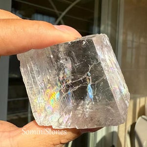 Puede incluir: Un cristal claro e iridiscente con forma rectangular. El cristal muestra una gama de colores, incluyendo azules, verdes y amarillos, debido a la refracción de la luz. El texto "SominiStones" es visible en la parte inferior. Es un espécimen mineral natural.