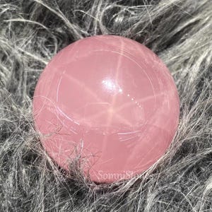 Puede incluir: Una esfera pulida de cuarzo rosa, de color rosa, con un patrón en forma de estrella. La esfera descansa sobre un lecho de material gris y esponjoso. El texto "SomniStones" es visible en la parte inferior derecha.