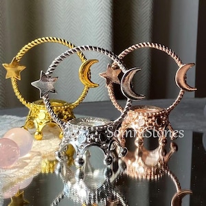 Puede incluir: Tres soportes decorativos de metal para esferas en oro, plata y oro rosa. Cada soporte presenta un diseño circular con un acento de estrella y media luna. Los soportes se exhiben sobre una superficie reflectante.