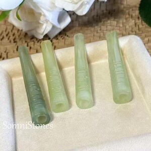 May include: Four pale green, jade-like, elongated objects with a tapered design and ridged detailing. The objects are arranged on a light-colored surface. The text "SomniStones" is visible in the lower left corner.