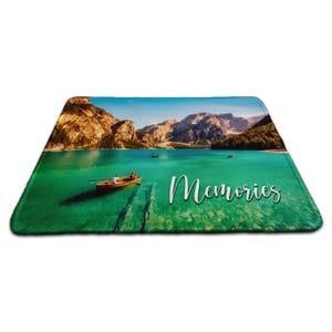 May include: Rectangular mat with a scenic lake view, a wooden boat, and mountains. The turquoise water reflects the mountains under a blue sky. The word "Memories" is written in white script.