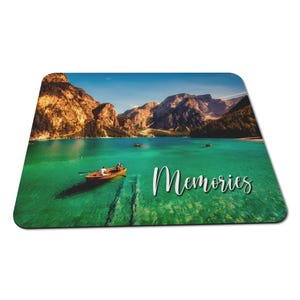 May include: Rectangular mouse mat with a scenic lake view, mountains, and a boat. The water is a vibrant turquoise, and the mountains are brown. The word "Memories" is written in white script across the image.
