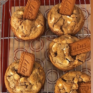 Op de afbeelding: Close-up van zes versgebakken koekjes met witte chocolade chips en een Lotus Biscoff koekje erop. De koekjes liggen op een metalen afkoelrek boven een rode siliconen bakmat.