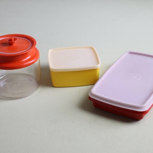 May include: Three vintage plastic food storage containers. A clear jar with an orange lid, a yellow square container with a cream lid, and a red rectangular container with a pink lid.