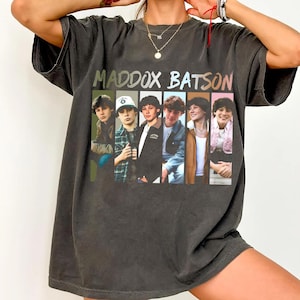 May include: Dark gray oversized t-shirt featuring the text "MADDOX BATSON" above a vertical row of six images of young men. The images are in a variety of outfits, including jackets, shirts, and a baseball cap. The shirt has short sleeves.