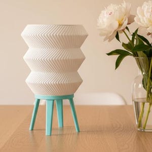 May include: A white, geometric vase with a chevron pattern, supported by a light blue tripod base. The vase is on a wooden table, next to a glass vase with white flowers. A framed abstract art piece hangs on the wall.