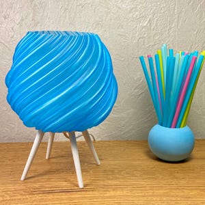 Może przedstawiać: Niebieska lampa z wirującym wzorem i białymi nogami oraz mały niebieski wazon wypełniony kolorowymi słomkami. Lampa ma unikalny, teksturowany design. Słomki są w różnych odcieniach niebieskiego, różowego i żółtego. Na drewnianej powierzchni.