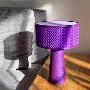 May include: A purple table lamp with a cylindrical shade and a conical base. The shade has a woven pattern, and the base has a glittery finish. The lamp is on a wooden surface.