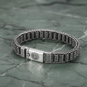 Puede incluir: Una pulsera de plata con un diseño de cadena trenzada. La pulsera presenta un cierre rectangular con un emblema grabado y cuentas cilíndricas decorativas con patrones grabados. La pulsera se muestra sobre una superficie de mármol verde.