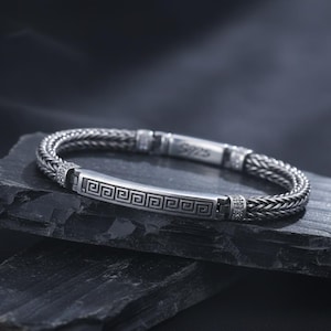 Könnte beinhalten: Ein silbernes Armband mit geflochtenem Band und einem rechteckigen Mittelstück mit griechischem Schlüsselmuster. Das Armband hat einen Verschluss und liegt auf einer dunklen, strukturierten Oberfläche.
