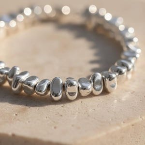 Könnte beinhalten: Ein kreisförmiges Armband, bestehend aus zahlreichen kleinen, polierten silberfarbenen Perlen mit unregelmäßigen, organischen, kieselsteinartigen Formen. Die Perlen sind zu einer durchgehenden Schlaufe aufgereiht und reflektieren das Licht auf ihren glatten, metallischen Oberflächen.