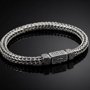 Op de afbeelding: Een zilveren armband met een gevlochten kettingontwerp. De armband heeft een rechthoekige sluiting met een gegraveerd patroon. De armband wordt gepresenteerd op een zwart oppervlak, dat het licht reflecteert.