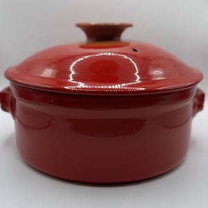 Puede incluir: Una cazuela de cerámica roja con tapa a juego. La tapa tiene un pequeño asa marrón y un orificio de ventilación. El plato tiene dos asas laterales. El acabado brillante refleja la luz. Un artículo de cocina.