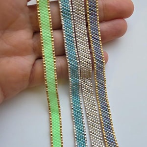 Pode incluir: Close-up de quatro pulseiras de contas em tons de verde, azul, prata e roxo. Cada pulseira tem aproximadamente 1,3 cm de largura e possui um fecho dourado. As pulseiras são seguradas em uma mão sobre um fundo branco.
