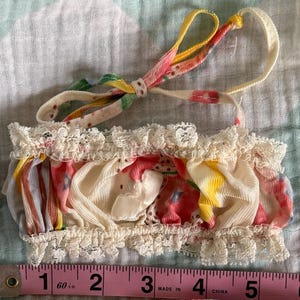 May include: A vintage two-piece baby outfit. The top is a ruffled crop top with a lace trim and a tie. The bottom is a pair of ruffled bloomers with a lace trim. The outfit features a floral pattern with pink, yellow, and blue accents. The outfit measures approximately 6 inches in length.