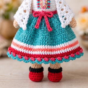 May include: A handmade crochet doll featuring a white bodice with colorful embroidery, a teal skirt with horizontal stripes in white, pink, and red, and a white lacy shawl. The doll wears red shoes with black cuffs and has a pink yarn bow at the waist.