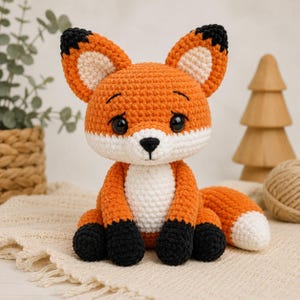 Op de afbeelding: Een gehaakte vossenknuffel in oranje, wit en zwart. De vos heeft grote, donkere ogen, een witte borst en zwarte poten. De oren zijn oranje met zwarte punten. De vos zit op een crèmekleurig oppervlak.