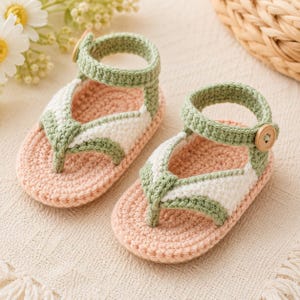 Könnte beinhalten: Ein Paar handgefertigte Baby-Häkelsandalen. Die Sandalen haben eine rosa Sohle, weiße und grüne Riemen und einen Verschluss mit Holzknopf. Die Sandalen im Zehensteg-Stil sind für ein Baby geeignet.