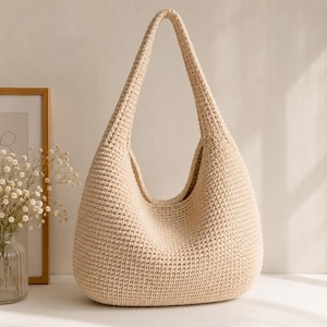 Op de afbeelding: Een beige gehaakte hobo-tas met een lange, gebogen handvat. De tas heeft een afgeronde bodem en een brede opening. De textuur van het haakwerk is zichtbaar en de tas lijkt gemaakt van een zachte, natuurlijke vezel. De tas is leeg en staat op een wit oppervlak.