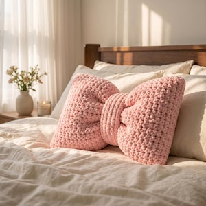 Könnte beinhalten: Ein hellrosa, schleifenförmiges Zierkissen. Das Kissen besteht aus einem grob gestrickten Material und liegt auf einem Bett mit weißer Bettwäsche. Die Mitte der Schleife ist eine dickere, zylindrische Form.