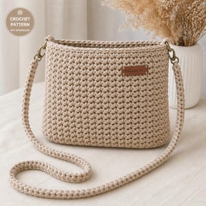 Peut inclure: Un sac à bandoulière en crochet beige de forme rectangulaire avec une longue sangle. Le sac présente un motif de point texturé et une petite étiquette en cuir marron avec l'inscription "handmade". Le sac est conçu avec du matériel métallique et est présenté sur une surface neutre.