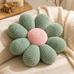 May include: A flower-shaped throw pillow featuring a central circular cushion in soft pink surrounded by eight rounded, plush petals in a muted sage green color. The entire item is constructed from a textured, crocheted yarn material.