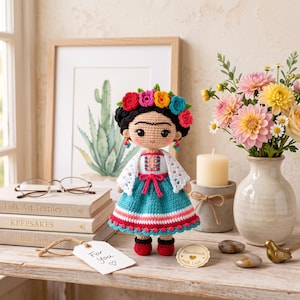 May include: A crocheted doll featuring dark hair styled with a colorful floral crown, wearing a white shawl over a teal skirt with red and white striped accents. The doll has black eyes, a small smile, and red shoes, standing upright on a wooden surface.
