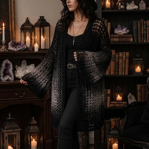 Op de afbeelding: Een lange, open gehaakte cardigan in een zwart-grijze ombre-design. De cardigan heeft wijde klokmouwen en een losse, open weefsel. Het model draagt een zwarte top en broek, met een zilveren ketting en een zwarte hoed.