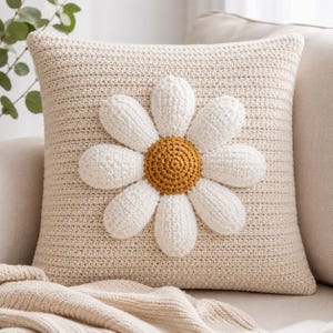 May include: Cream-colored crochet pillow with a large, white daisy applique. The daisy has a golden-yellow center. The pillow is square and appears to be made of a soft, textured yarn, perfect for home decor.