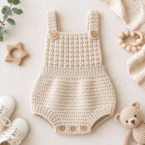 Puede incluir: Un pelele de bebé de crochet color crema con cierres de botones de madera. El pelele presenta un patrón de puntadas texturizadas y tirantes ajustables. Artículos adicionales incluyen un mordedor de madera en forma de estrella, zapatos de bebé blancos y un pequeño osito de peluche de ganchillo.