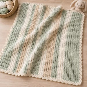 May include: A handmade crochet baby blanket with vertical stripes in soft green, cream, and beige. The blanket has a scalloped edge and is made of a soft yarn. A basket of yarn and a stuffed bunny are in the background.