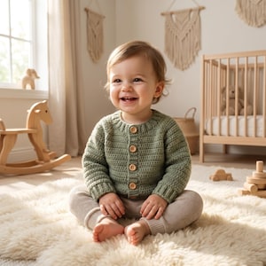 May include: A baby wearing a sage green crochet cardigan with wooden buttons and beige trousers, smiling in a nursery. The baby sits on a fluffy white rug. A wooden rocking horse and cot are visible in the background.