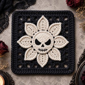 May include: A square, black crocheted item featuring a cream-colored skull design with a floral arrangement. The skull has black eyes and a stitched mouth. The square has a textured, openwork border. The item is likely a decorative piece or part of a larger craft project.