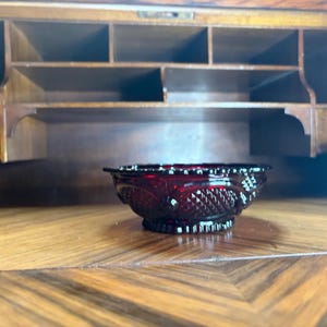 May include: A small, ruby-red glass bowl with a detailed, textured pattern. The bowl is set on a wooden surface with a geometric design, with a wooden cabinet in the background. The bowl's design includes a diamond pattern.