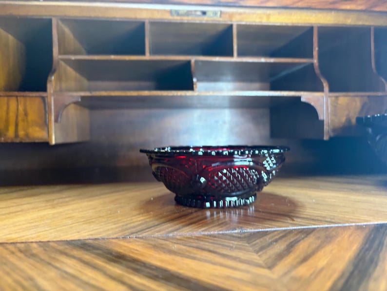 May include: A small, ruby-red glass bowl with a detailed, textured pattern. The bowl is set on a wooden surface with a geometric design, with a wooden cabinet in the background. The bowl's design includes a diamond pattern.