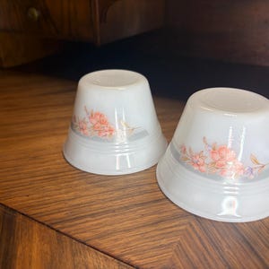 May include: Two white glass thimbles with a floral design in shades of pink, orange, and grey. The thimbles have a flared shape and are set against a wooden surface.