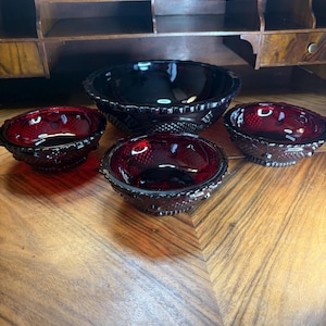 May include: Four ruby red glass bowls with textured exteriors. The bowls are of varying sizes, with the largest in the centre and three smaller ones around it. The bowls are displayed on a wooden surface. The bowls are likely vintage.